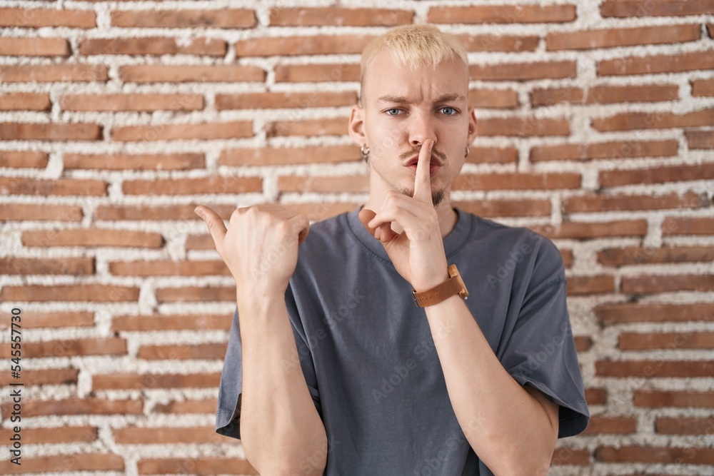 Young caucasian man standing over bricks wall asking to be quiet with finger on lips pointing with hand to the side. silence and secret concept.