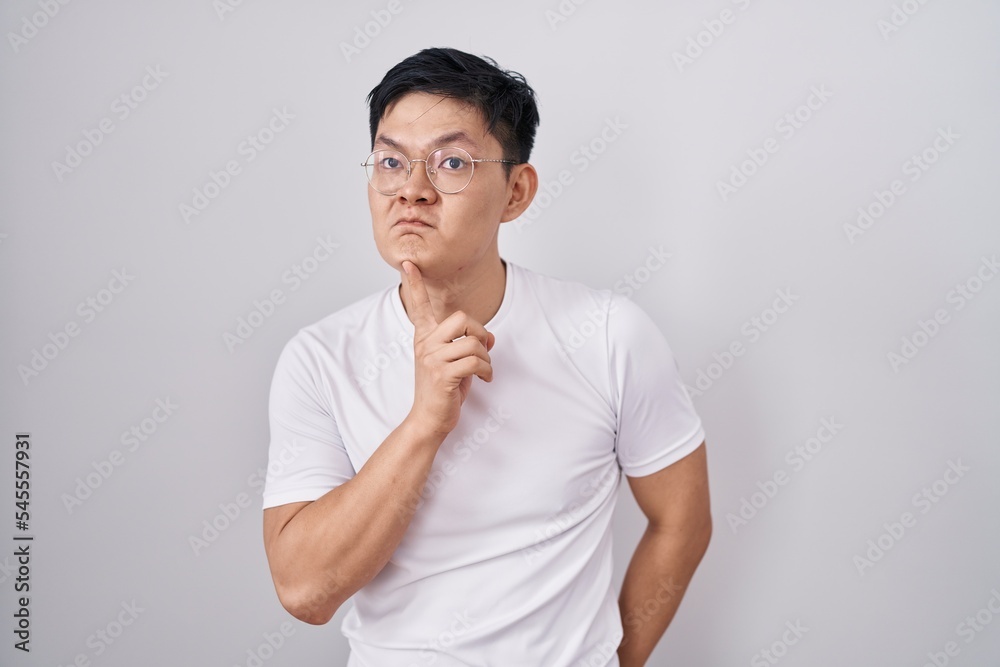 Young asian man standing over white background thinking concentrated about doubt with finger on chin and looking up wondering Young asian man standing over white background thinking concentrated about doubt with finger on chin and looking up wondering