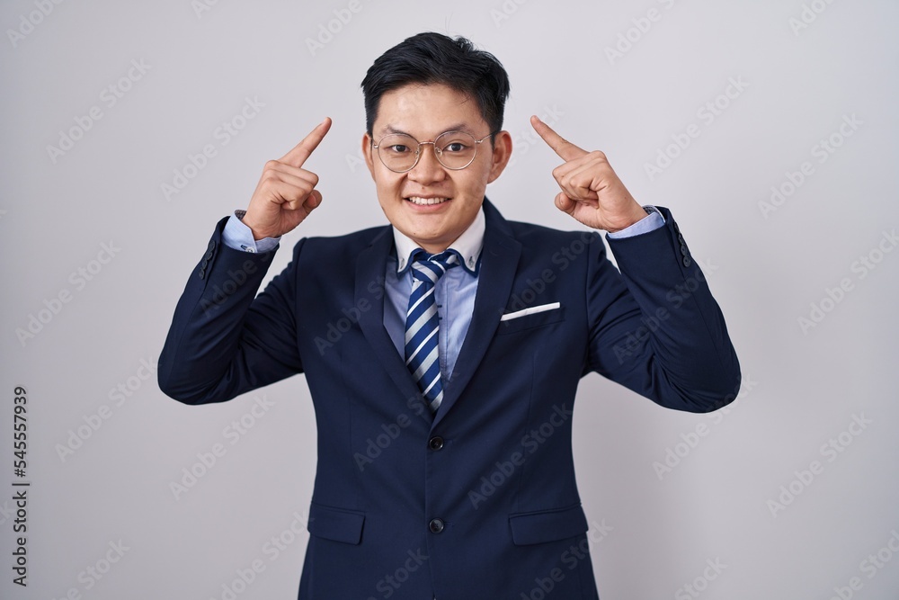 Young asian man wearing business suit and tie smiling pointing to head with both hands finger, great idea or thought, good memory Young asian man wearing business suit and tie smiling pointing to head with both hands finger, great idea or thought, good memory