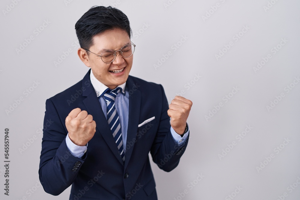 Young asian man wearing business suit and tie very happy and excited doing winner gesture with arms raised, smiling and screaming for success. celebration concept. Young asian man wearing business suit and tie very happy and excited doing winner gesture with arms raised, smiling and screaming for success. celebration concept.