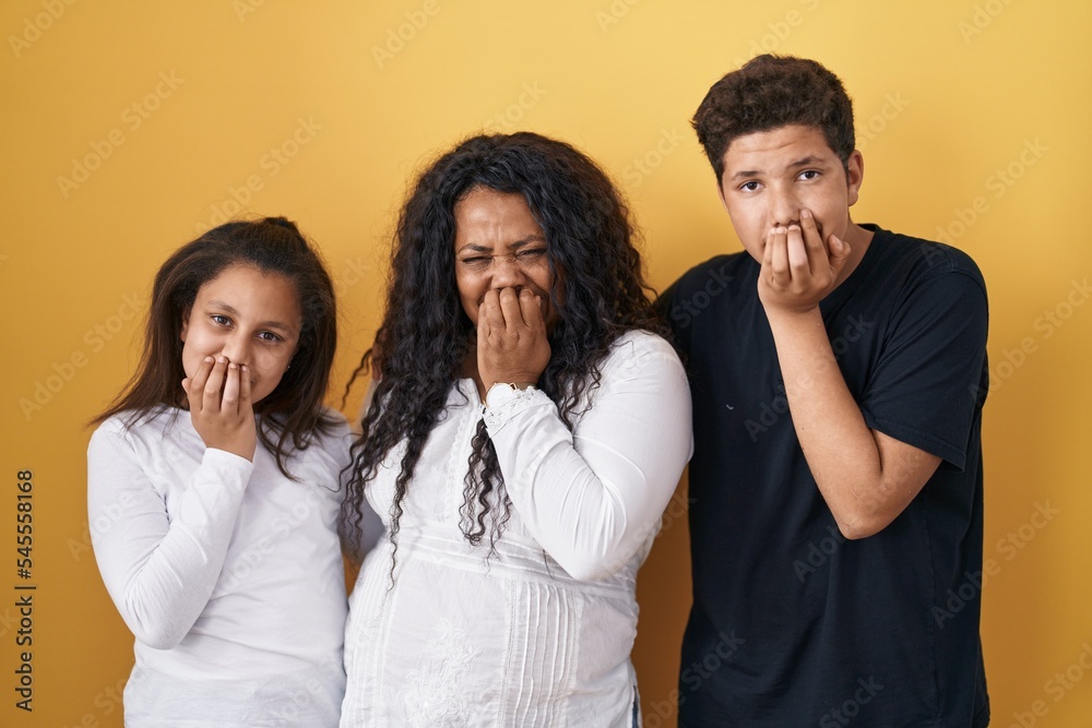 Family of mother, daughter and son standing over yellow background laughing and embarrassed giggle covering mouth with hands, gossip and scandal concept Family of mother, daughter and son standing over yellow background laughing and embarrassed giggle covering mouth with hands, gossip and scandal concept