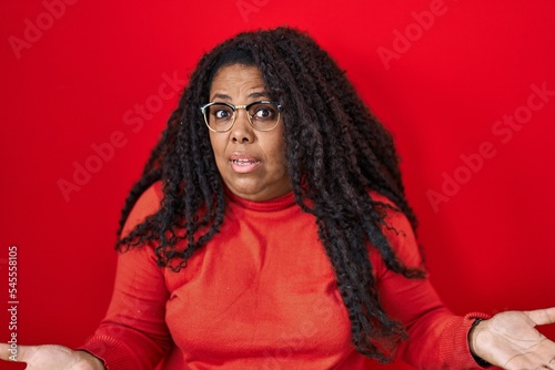 Plus size hispanic woman standing over red background clueless and confused with open arms, no idea concept. © Krakenimages.com