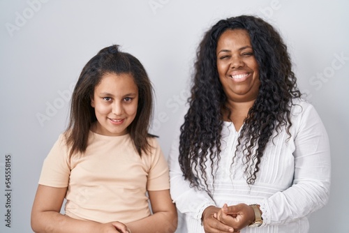 Mother and young daughter standing over white background with hands together and crossed fingers smiling relaxed and cheerful. success and optimistic