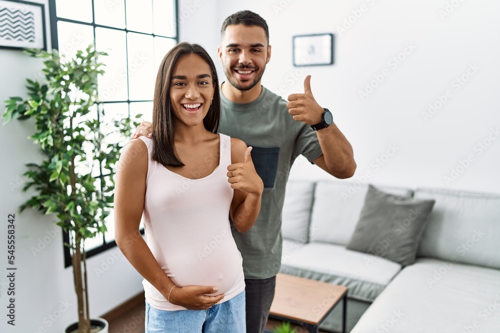 Young interracial couple expecting a baby, touching pregnant belly doing happy thumbs up gesture with hand. approving expression looking at the camera showing success.