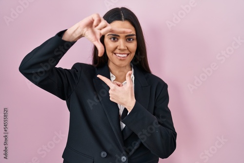Young brunette woman wearing business style over pink background smiling making frame with hands and fingers with happy face. creativity and photography concept.
