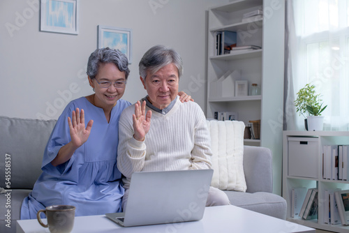 Happy old Asian senior grandparents couple waving hand making online video call enjoying family virtual meeting digital webcam videocall chat talking  looking at laptop computer on couch at home.