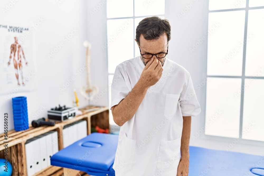 Middle age man with beard working at pain recovery clinic tired rubbing nose and eyes feeling fatigue and headache. stress and frustration concept. Middle age man with beard working at pain recovery clinic tired rubbing nose and eyes feeling fatigue and headache. stress and frustration concept.