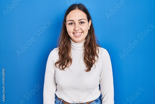 Young hispanic woman standing over blue background with a happy and cool smile on face. lucky person.