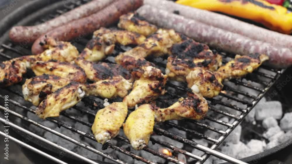 Delicious and appetizing grilled chicken wings with sausages and peppers being cooked on the grill in the backyard of the house, close-up side view in slow motion with zoom out. Delicious and appetizing grilled chicken wings with sausages and peppers being cooked on the grill in the backyard of the house, close-up side view in slow motion with zoom out.