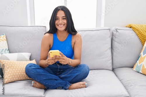 Young hispanic woman using smartphone sitting on sofa at home