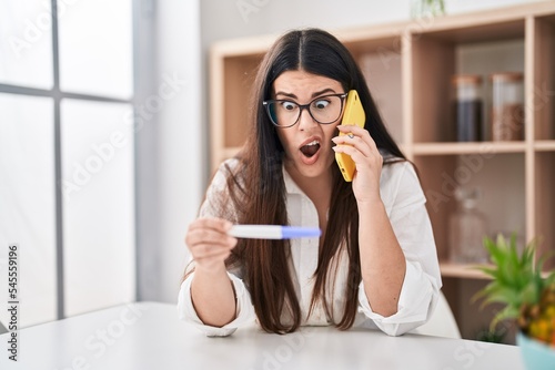 Young brunette woman holding pregnancy test result speaking on the phone clueless and confused expression. doubt concept.