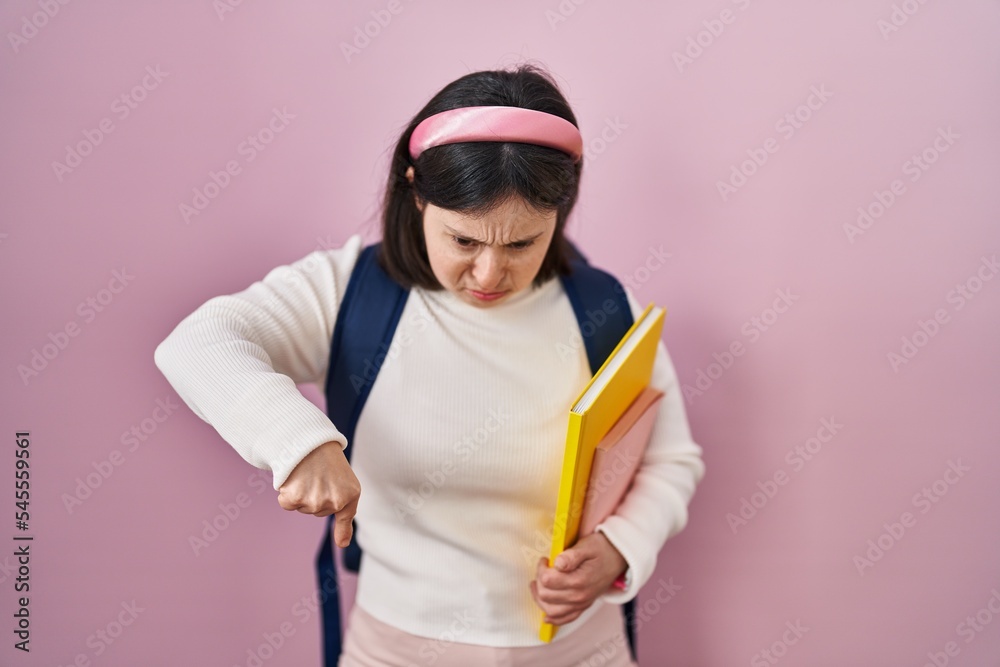 Woman with down syndrome wearing student backpack and holding books pointing down looking sad and upset, indicating direction with fingers, unhappy and depressed. Woman with down syndrome wearing student backpack and holding books pointing down looking sad and upset, indicating direction with fingers, unhappy and depressed.