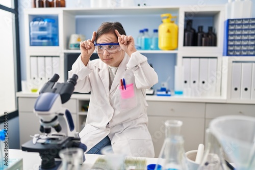 Hispanic girl with down syndrome working at scientist laboratory doing funny gesture with finger over head as bull horns