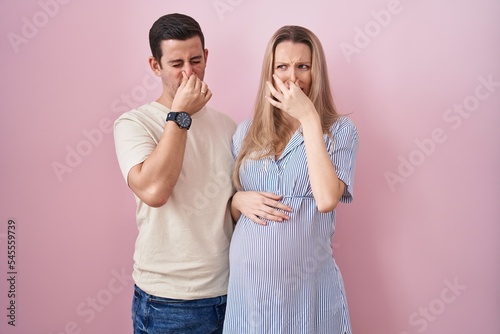 Young couple expecting a baby standing over pink background smelling something stinky and disgusting, intolerable smell, holding breath with fingers on nose. bad smell
