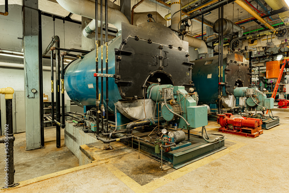 The interior of an old and well used boiler room with two large boilers, many pipes, valves and other equipment.
