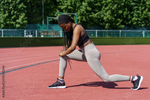 Sportive young woman with long braids takes care of body on summer day. African American lady in sportswear enjoys effective exercise for gluteal muscles on city stadium near park with green trees