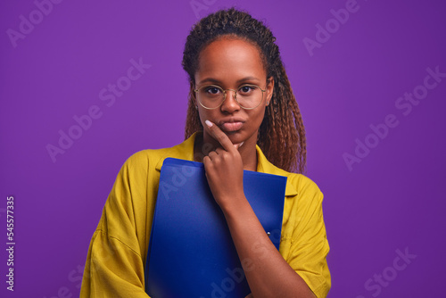 Young capricious casual African American woman pout touches chin and presses file folder to chest after not liked conversation or remark from manager stands posing in purple studio. Ethnic girl