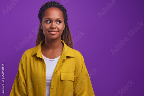 Young cute positive African American woman looks away with shy smile when sees likes person or interesting product with good discount on purchase standing posing on purple background. Copy space