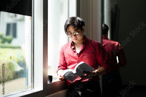 Learning form reading, Portrait of man wear red shirt and glasses looking smart reading book standing beside window and cup of coffee with high contrast lighting