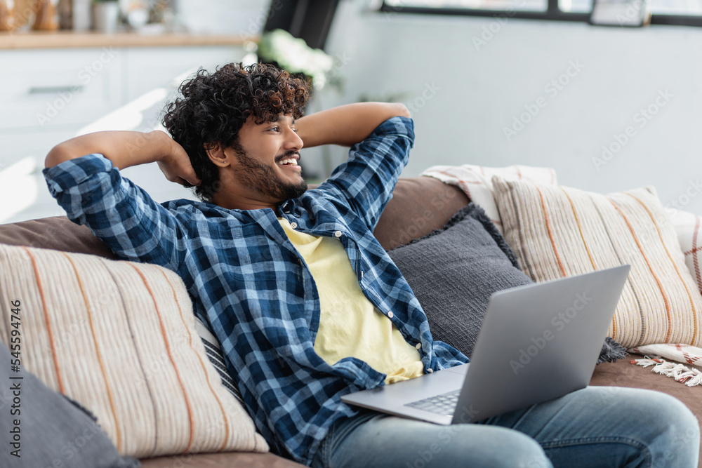 Fototapeta premium Young Indian freelance man with laptop throwing his hands behind his head distracted from work sitting on the couch, looking away while, thinking about something positive and smiling