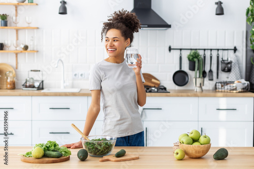 Woman cooking healthy food with glass of clean water, looking away and smiling. African American young female preparing fresh salad for dinner or breakfast in home kitchen. Healthy diet eating photo