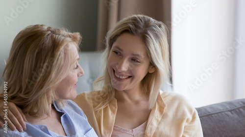 Woman hugs 50s mom talking seated on sofa. Mature female enjoy communication to young adult daughter  family lead trustworthy conversation relaxing  spend leisure at home. Bond  understanding  support