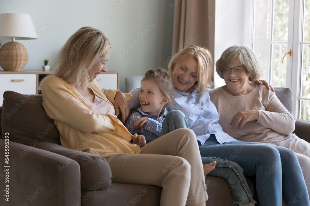 Little girl spend time with loving mom, grandmother and elderly great-grandmother sit together on sofa laughing play feel happy. Multi-generational women enjoy communication, having friendly relations