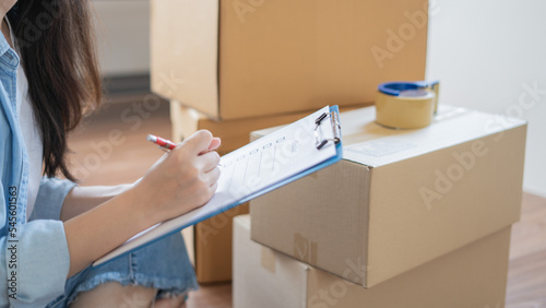 Asian woman checking and packing the carton box prepare to move to new house relocation shipping or check goods package before delivery to customer from online shopping by checklist