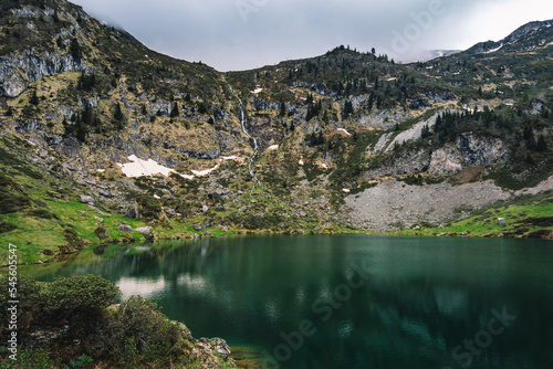 Fototapeta Naklejka Na Ścianę i Meble -  View on the beautiful blue lake of Ayes, with a waterfall, the pastures and the peaks of the French Pyrenees on a cloudy day in the background