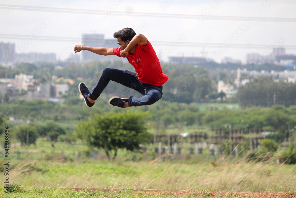 An energetic young man in mid-air performs a martial arts-style jump. Dressed in red and dark pants, he strikes a powerful pose against a grassy field and blurred cityscape, symbolizing energy. An energetic young man in mid-air performs a martial arts-style jump. Dressed in red and dark pants, he strikes a powerful pose against a grassy field and blurred cityscape, symbolizing energy.