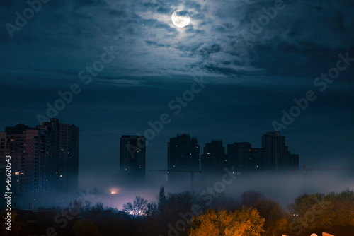Night cityscape. The full moon through the clouds over city street at night with high modern buildings among the fog with illumination from a street light. Blackout in Kyiv. Ukraine.