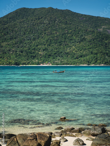Scenic view of Sunset Beach, Koh Lipe Island. Small local long tail boat on crystal clear turquoise sea with coral reef transparent shallow water against Koh Adang Island background. Satun, Thailand.