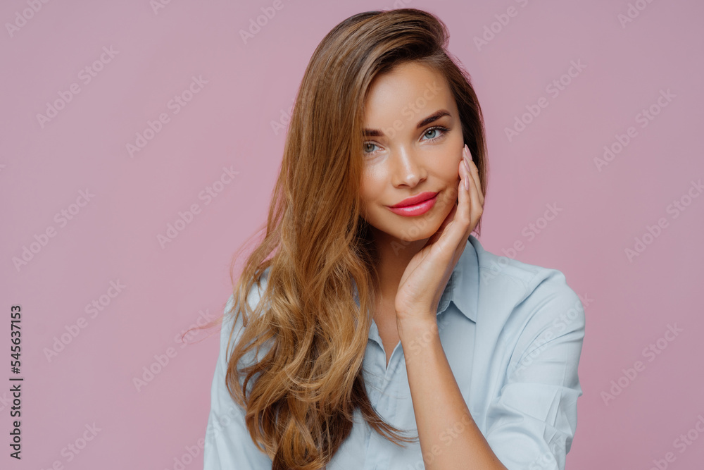 Studio shot of pleasant looking relaxed female model with long hair touches cheek gently, wears elegant shirt, looks straightly at camera, shows her beauty. Pretty businesswoman prepares to work Studio shot of pleasant looking relaxed female model with long hair touches cheek gently, wears elegant shirt, looks straightly at camera, shows her beauty. Pretty businesswoman prepares to work
