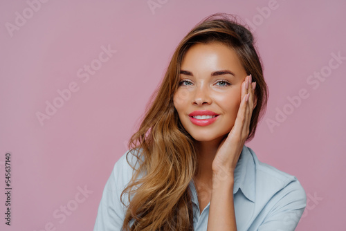 Photo of pleasant looking female model touches cheeks with palm, has gentle toothy smile, wears makeup, dressed in casual shirt, stands against purple wall, copy space area for your advertisement