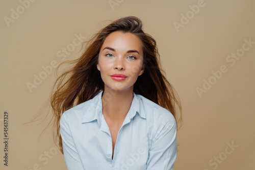 Lovely young female has long wavy hair floating in wind, dressed in elegant blue shirt, wears lipstick and makeup, poses against brown background, has direct gaze at camera, healthy facial skin