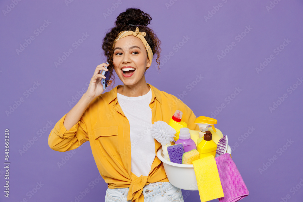 Young smiling housekeeper woman wear yellow shirt hold basin with detergent bottles talk speak on mobile cell phone tidy up isolated on plain pastel light purple background studio. Housework concept. Young smiling housekeeper woman wear yellow shirt hold basin with detergent bottles talk speak on mobile cell phone tidy up isolated on plain pastel light purple background studio. Housework concept.