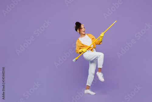 Full body side view fun young housekeeper woman wear yellow shirt tidy up hold broom sweeping brush look aside clean house isolated on plain pastel light purple background studio. Housework concept.