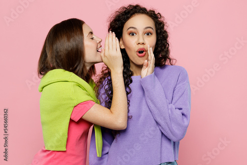 Fototapeta Young two friends shocked women wears green purple shirts together whispering gossip and tells secret behind her hand sharing news isolated on pastel plain light pink color background studio portrait