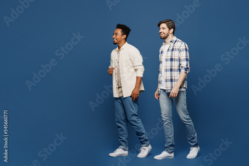 Full body side view young two friends smiling cheerful happy men 20s wear white casual shirts together walking going strolling isolated plain dark royal navy blue background. People lifestyle concept.