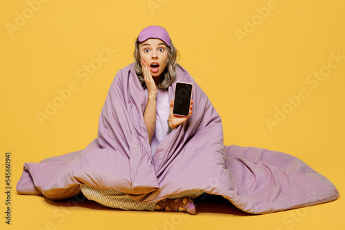 Full body young woman in purple pyjamas jam sleep eye mask rest relax at home sit wrap covered blanket duvet hold mobile cell phone blank screen isolated on plain yellow background Night nap concept © ViDi Studio