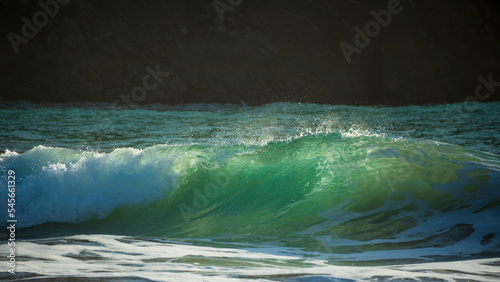 Majestic landscape image of jade turquoise waves crashing onto shore and rocks in Kynance Cove Cornwall with glowing sunrise background and water spray dorplets in wind