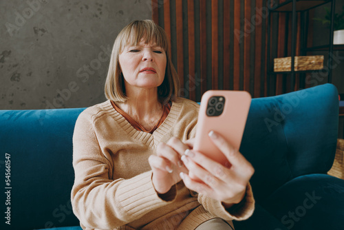 Elderly woman 50s years old wears casual clothes sits on blue sofa hold use mobile cell phone without glasses squint stay at home flat rest relax spend free spare time in living room indoor grey wall. photo