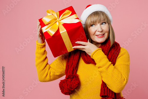 Merry elderly woman 50s years old wear knitted sweater red scarf Santa hat posing hold present box ribbon bowgift look aside isolated on plain pink background. Happy New Year Christmas 2023 concept.