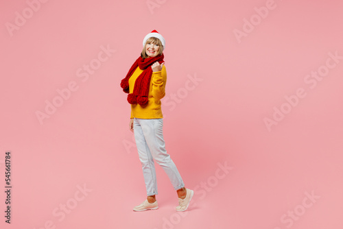 Full body side view elderly woman 50s year old wear yellow knitted sweater red scarf Santa hat posing point finger aside on area isolated on plain pink background Happy New Year Christmas concept
