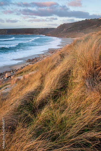 Stunning landscape image of Sennen Cove in Cornwall during sunset viewed from grassy sand dunes with dramatic sky and long exposure sea motion