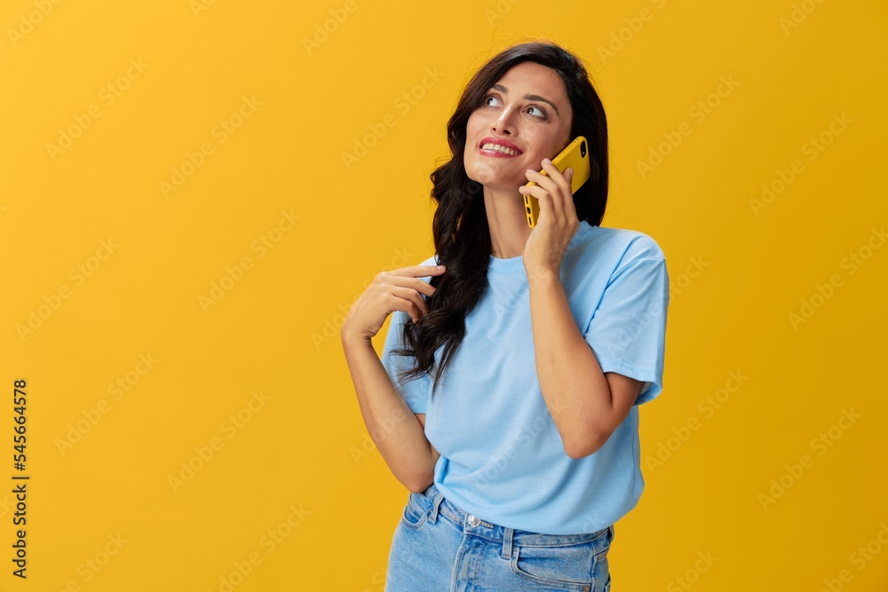 Woman blogger with a phone in her hands in a blue t-shirt and jeans on a yellow background smile signs gestures symbols, online communication and video call, copy space, free background