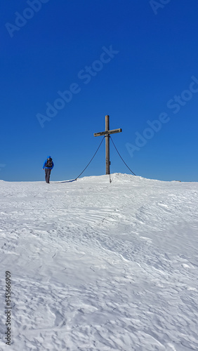 Man with hiking backpack walking in deep snow to summit cross of mountain peak Ladinger Spitz, Saualpe, Lavanttal Alps, Carinthia, Austria, Europe. Ski touring in Austrian Alps on sunny winter day