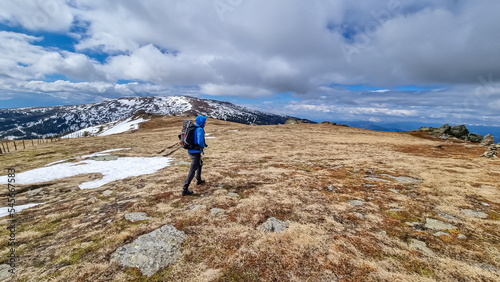 Rear view of man with hiking backpack walking on alpine meadow from Ladinger Spitz to Gertrusk, Saualpe, Lavanttal Alps, Carinthia, Austria, Europe. Trekking on cloudy early spring day. Wanderlust