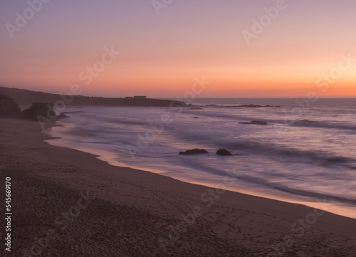 Mysterious long exposure view of sand beach Praia Grande de Almograve with blurred ocean waves in pink orange and purple light. Blue hour after sunset at Rota Vicentina coast, Almograve, Portugal