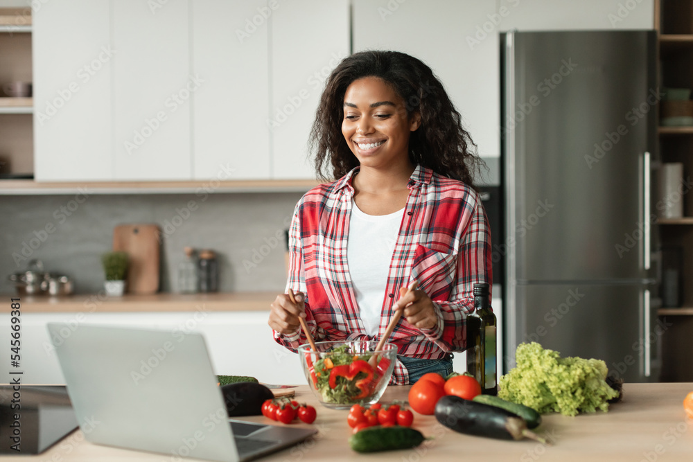 Glad black young lady in casual prepares salad from organic vegetables and watch video lesson of recipe